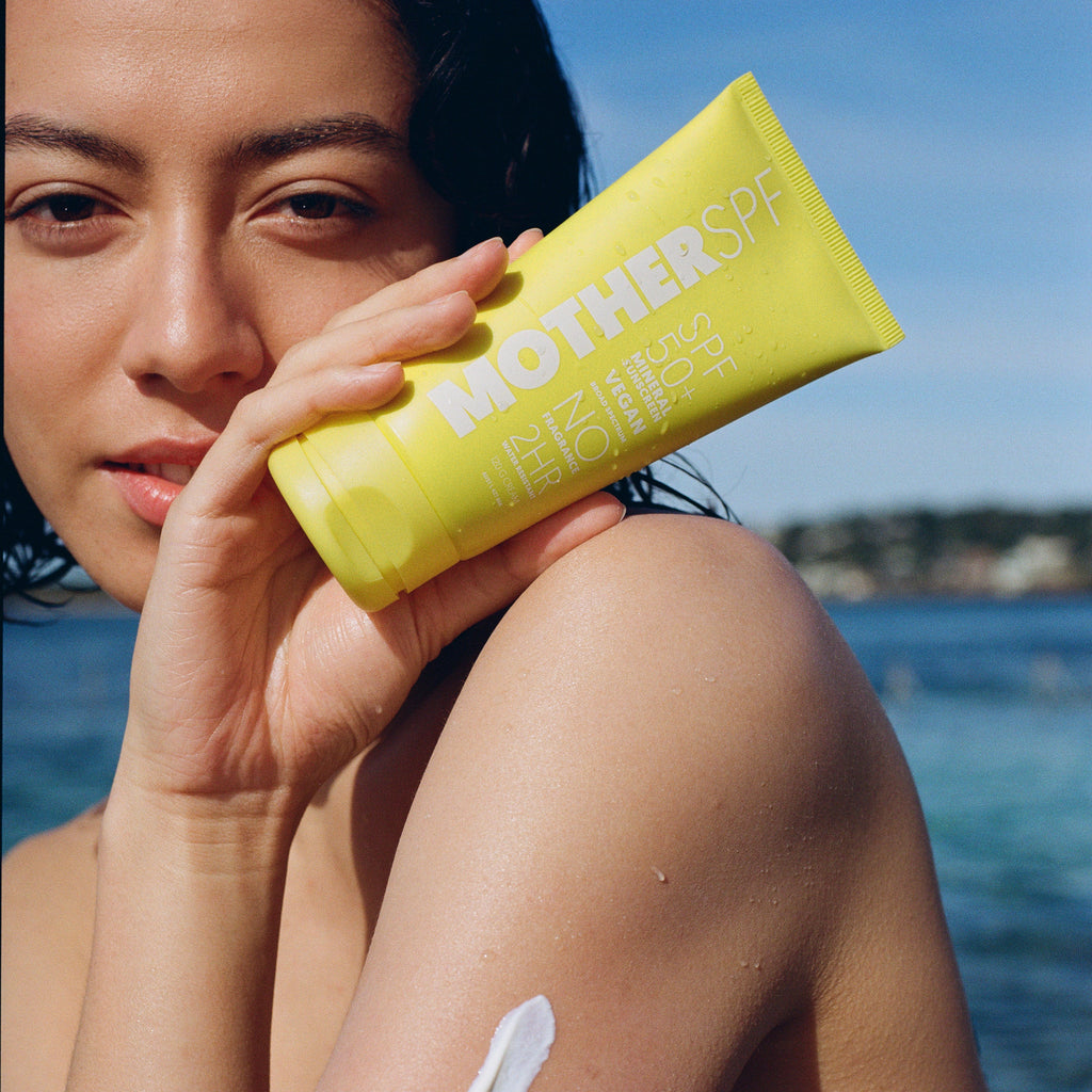 Woman applying Mother SPF 50 Face & Body holding yellow 'Mother SPF 50 Face & Body product by the beach.